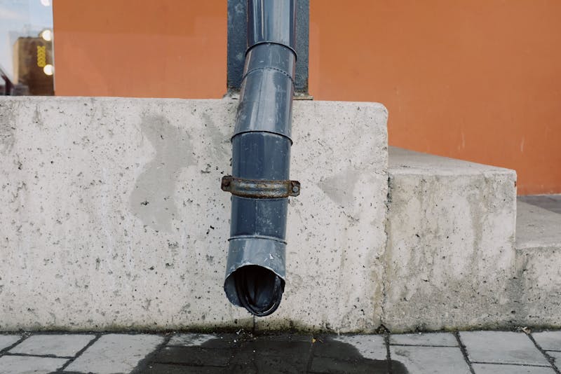 Downspout Cleaning — Aged metal drain pipe on a concrete wall with water stains, highlighting urban decay.